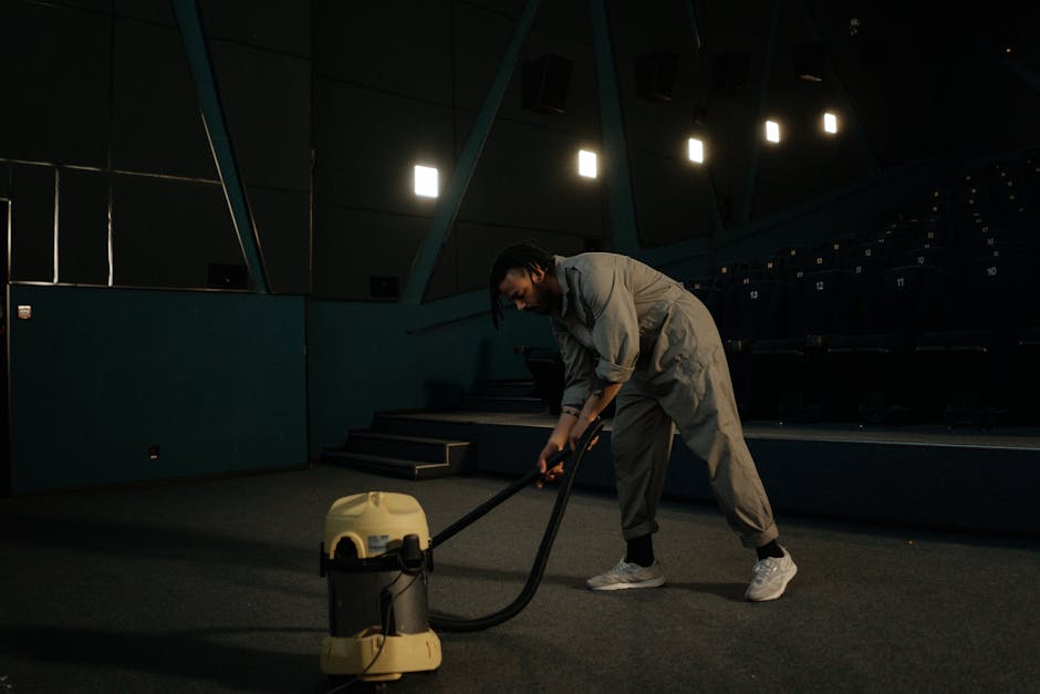 A person using a vacuum cleaner with a yellow and black body and a long black hose on an ornate, patterned area rug in a residential living room. The rug features floral motifs in muted colors, and th