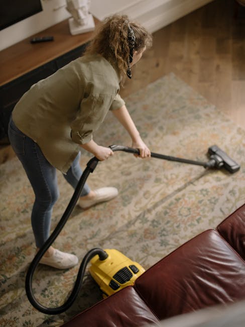A person kneeling on a patterned carpet in a room, using a yellow vacuum cleaner with a black hose for surface cleaning. The individual, dressed in a beige jacket and blue jeans, is reaching into the 