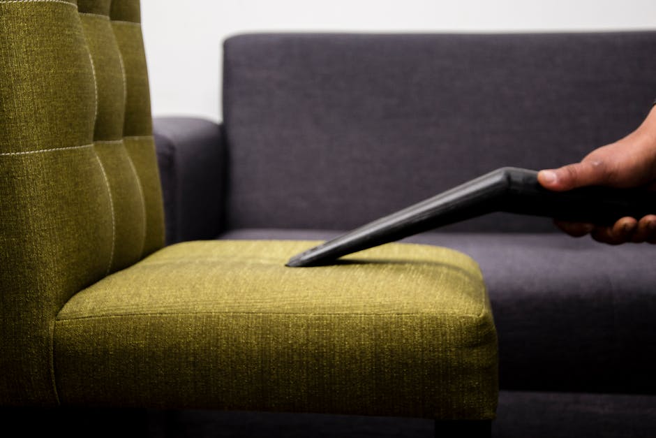 Close-up of a person's hand using a handheld vacuum cleaner to clean a green fabric upholstered chair in a living room setting. The chair has a tufted backrest with visible stitching and a smooth, wel